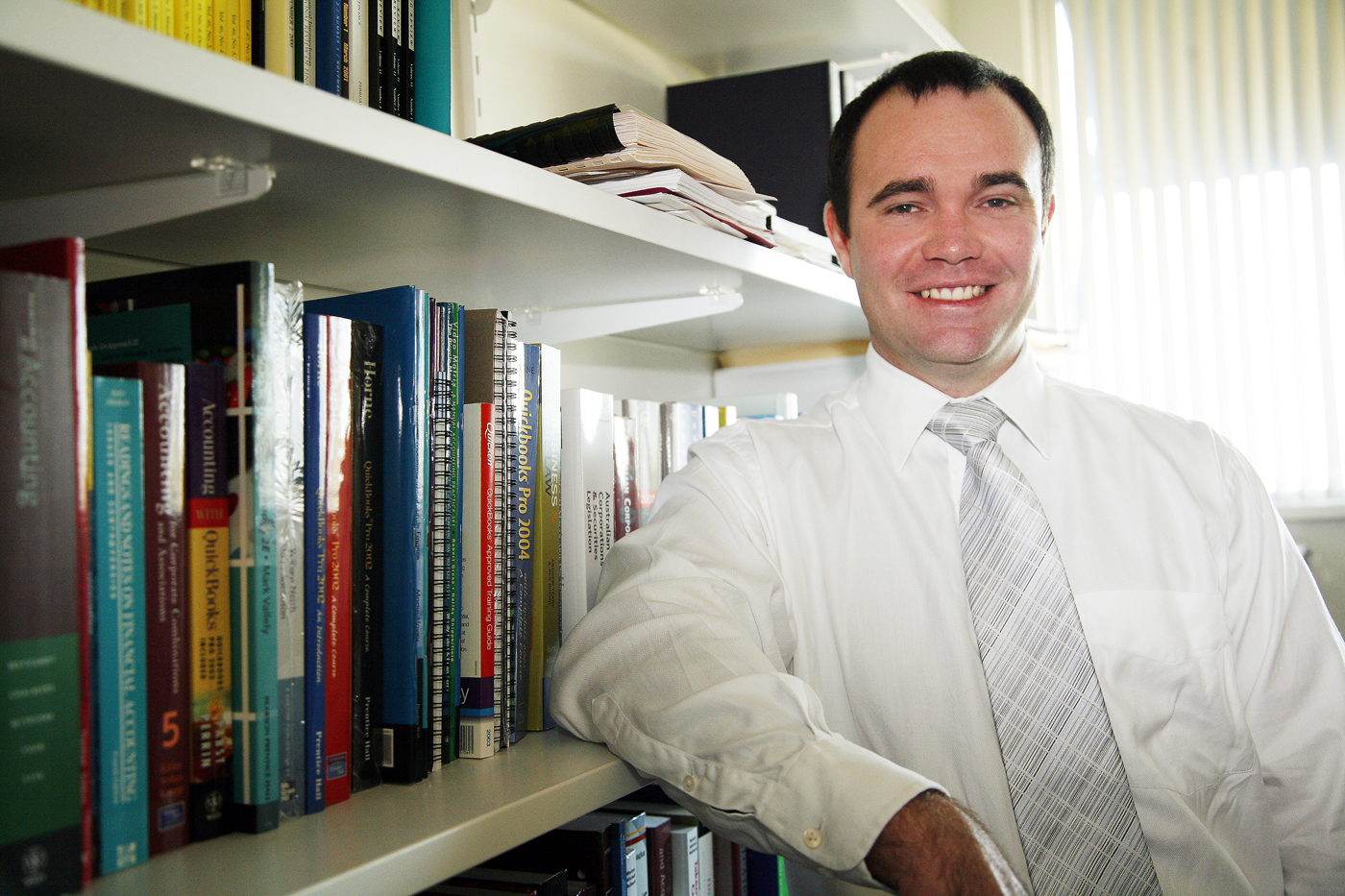 Mark Brimble, in shirt and tie, in office with arm on book shelf.