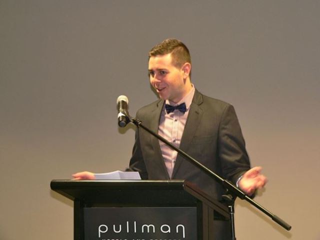 Matthew Littlejohn, dressed in tuxedo, at lectern, delivering speech.