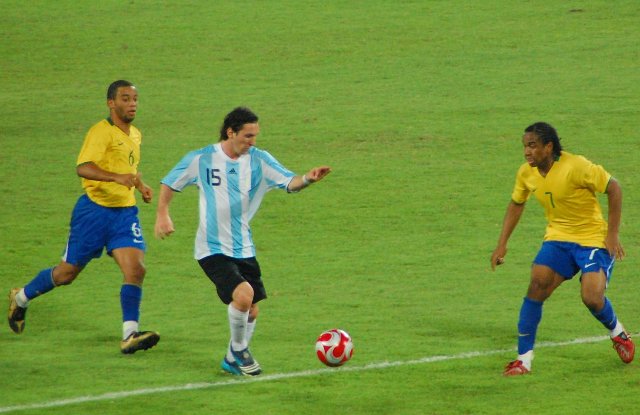 Three footballers, two in Brazil outfit, one in Argentina gear, competing for ball.