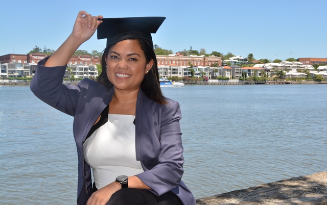 Griffith graduate Kathleen, dressed smartly, holds onto a graduating mortar board hat on her head with Brisbane river in background.