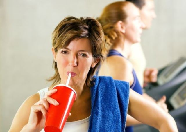 Women in sports outfit, towel on shoulder, drinking water through a straw from a red cup.