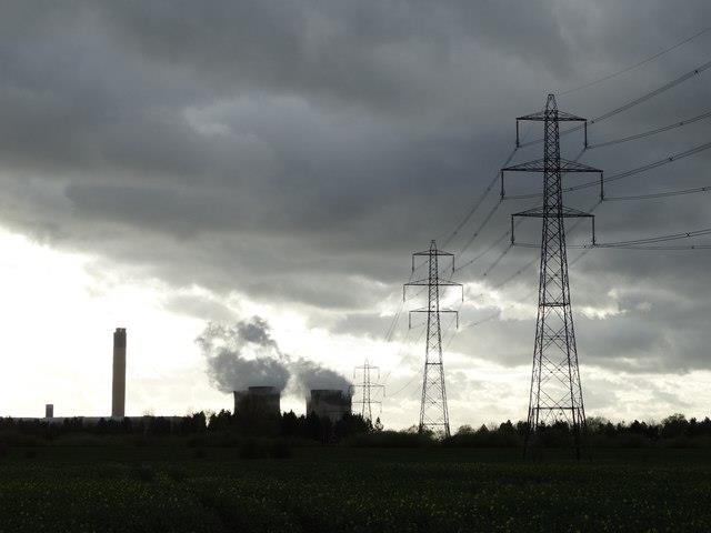 Black and white shot of pylons in foreground, clouds in sky and shadow of towers in background with smoke rising out.