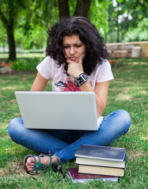 Woman sitting cross-legged in a park looking a laptop with some books on the grass.