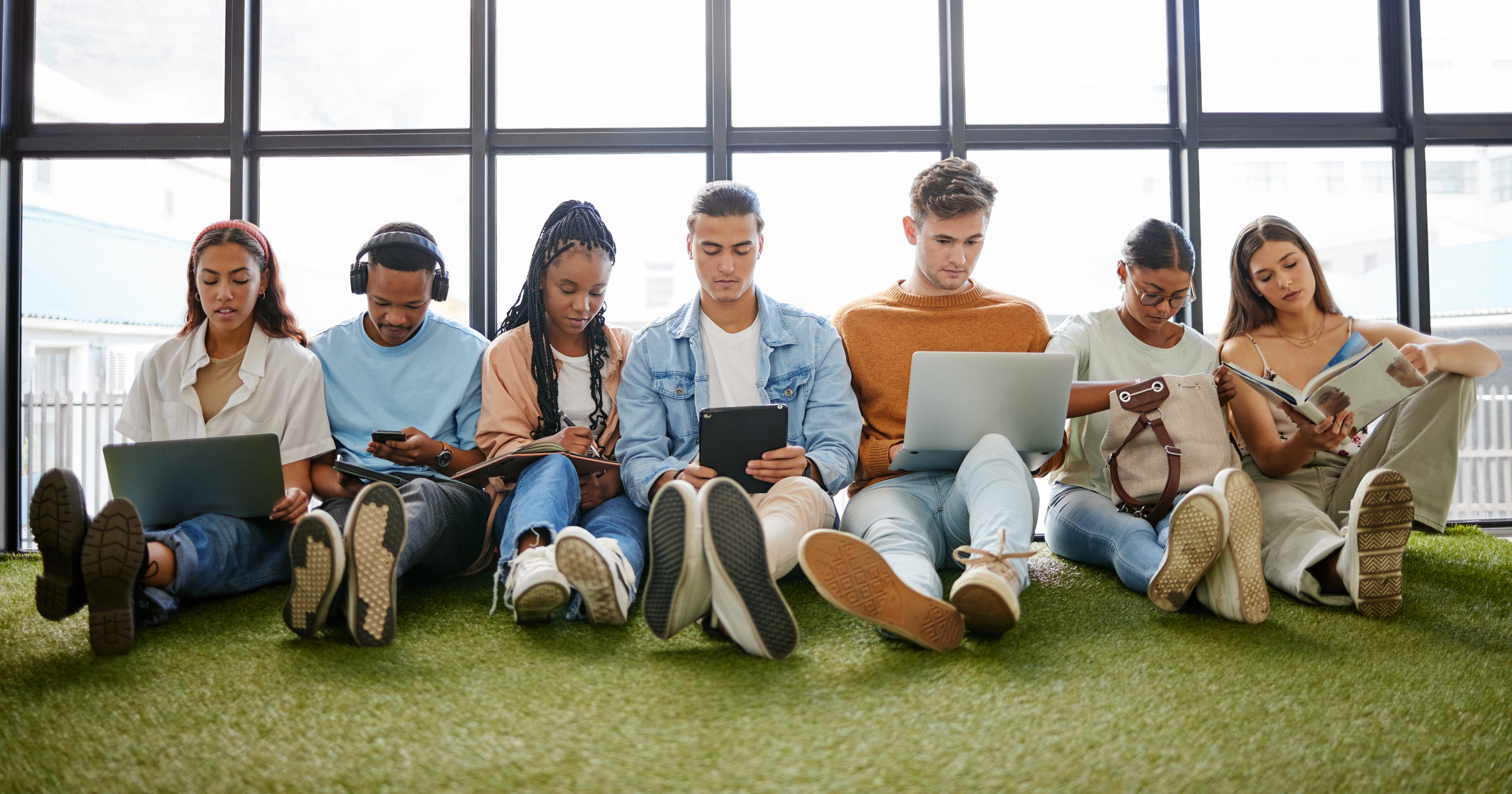 A line of young people sitting against a window. They are working on laptops and mobile phones.