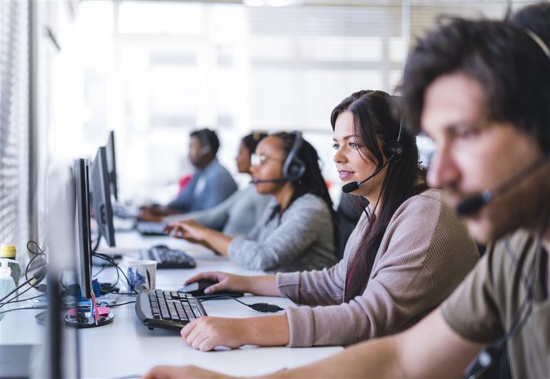 Call centre workers seated in a line, talking to callers with headsets and looking at a computer screen.